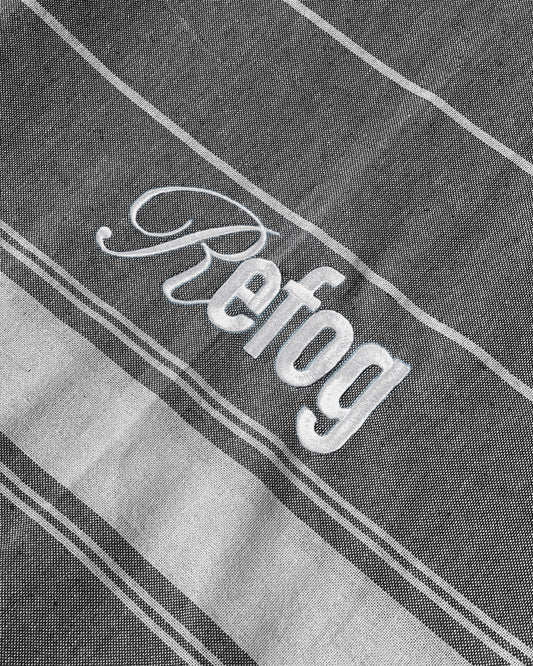 Detalle del logo bordado de Refog en toalla gris oscuro con rayas blancas